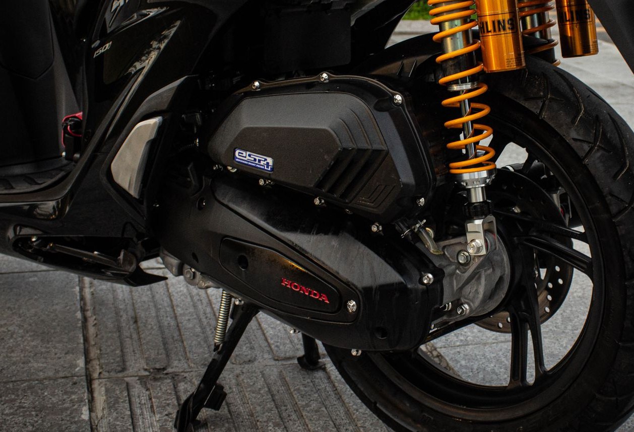 スクーターの左側面の寄り。後輪の前に、HONDAのロゴが入った黒く細長い駆動系のケースがあり、その後ろに黄色いばねのリアサスペンションと後輪が見える