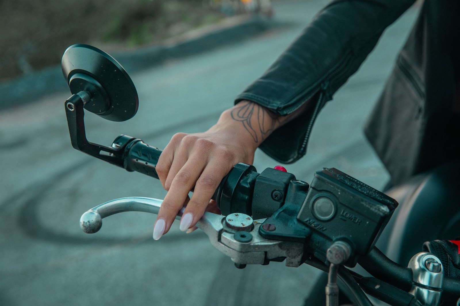 バイクの右ハンドルのグリップを握り、銀色のブレーキレバーに指をかけている手元。レバーの付け根にブレーキのマスターシリンダー、グリップの端に丸いミラーが付いている