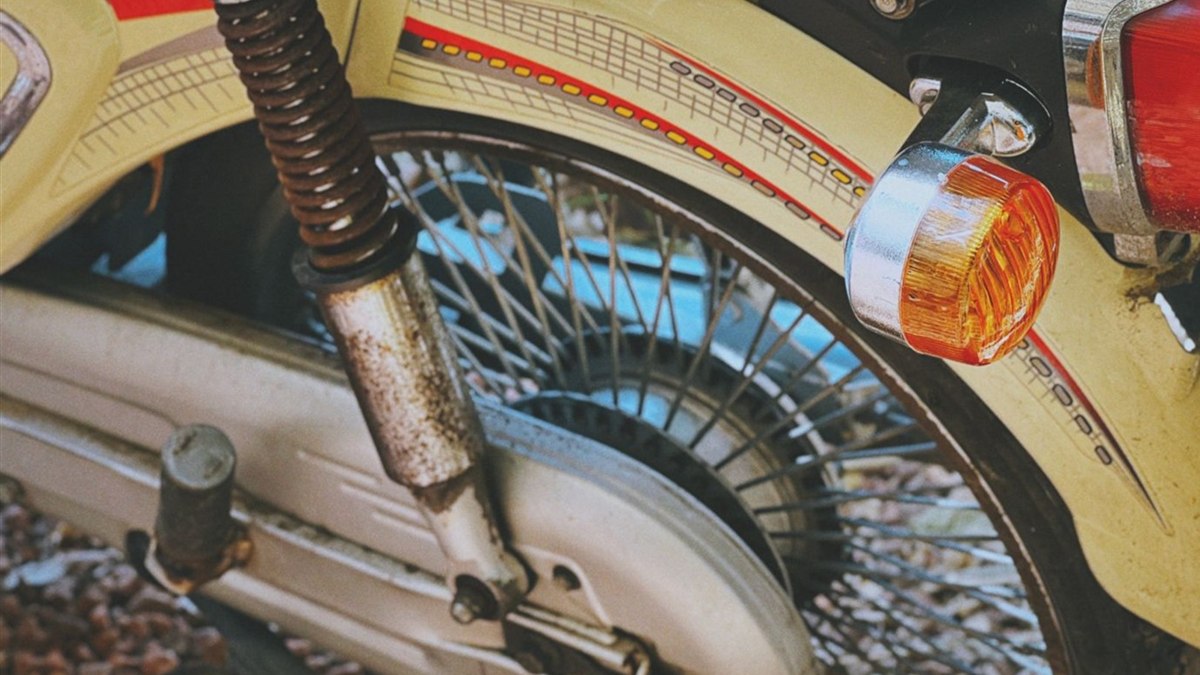 バイクの後輪まわりのクローズアップ。リアサスペンションのばねが茶色くサビ、銀色の筒に細かい点サビが出ている。奥にスポークホイール、右にオレンジのウインカーが見える