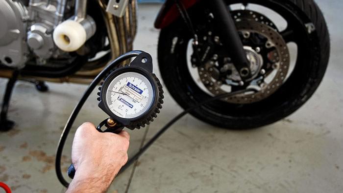 手に持った、ホースの付いたダイヤル式のタイヤゲージ。奥にバイクの前輪とディスクブレーキが写っている